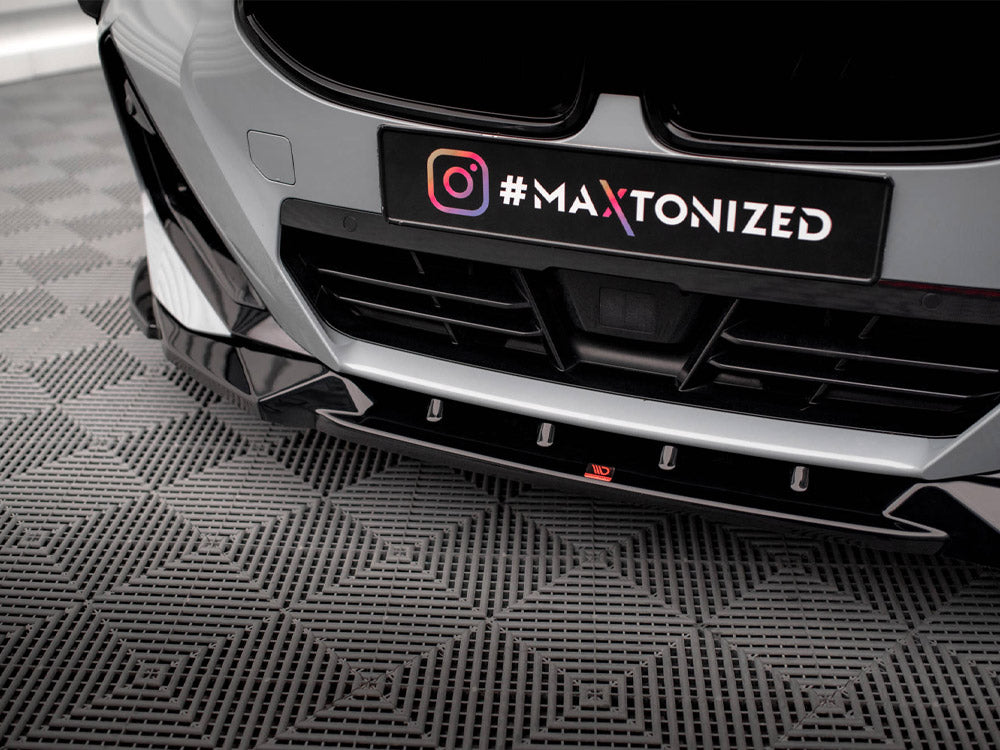 A car's front bumper with a black license plate holder, displaying "#MAXTONIZED" next to an Instagram logo, is parked on a textured gray floor.