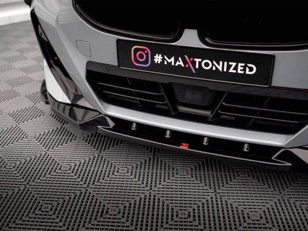 A car's front bumper with a black license plate holder, displaying "#MAXTONIZED" next to an Instagram logo, is parked on a textured gray floor.