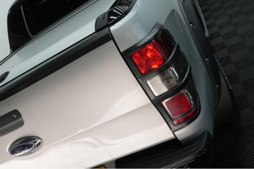 A white pickup truck with illuminated right rear taillights is parked on a dark, textured surface. A Ford logo is visible on the tailgate.