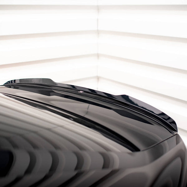 A sleek black car roof with a subtle spoiler is angled in a well-lit, white-striped room, casting dynamic reflections across its surface.