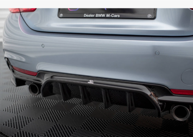 Car rear bumper with dual exhaust pipes and a black diffuser, parked on a textured surface. The license plate reads, "Dealer BMW M-Cars."