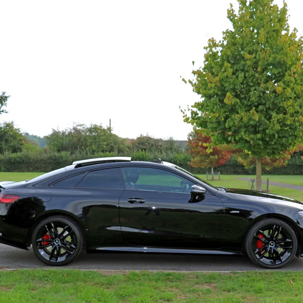 A black sports car is parked on a paved road, set against a grassy park landscape with trees in varying stages of seasonal color change.