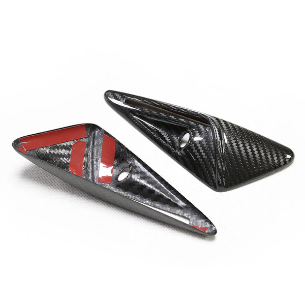 Two carbon fiber arrowhead-shaped objects, one featuring red adhesive strips, rest on a white surface. Each has a small circular hole, suggesting mounting capabilities.