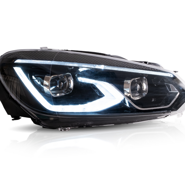 A car headlight displaying illuminated LED lights is positioned in a bright studio setting with reflections visible on its clear surface.