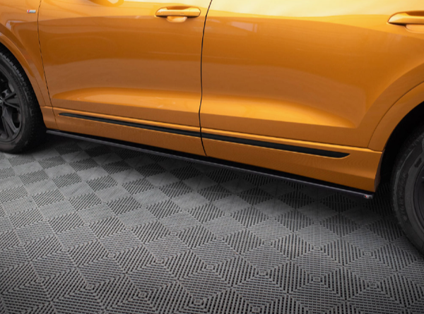A bright orange car is parked on a floor with a geometric, checkered pattern, showing part of the side, including the door and handle against the textured gray surface.