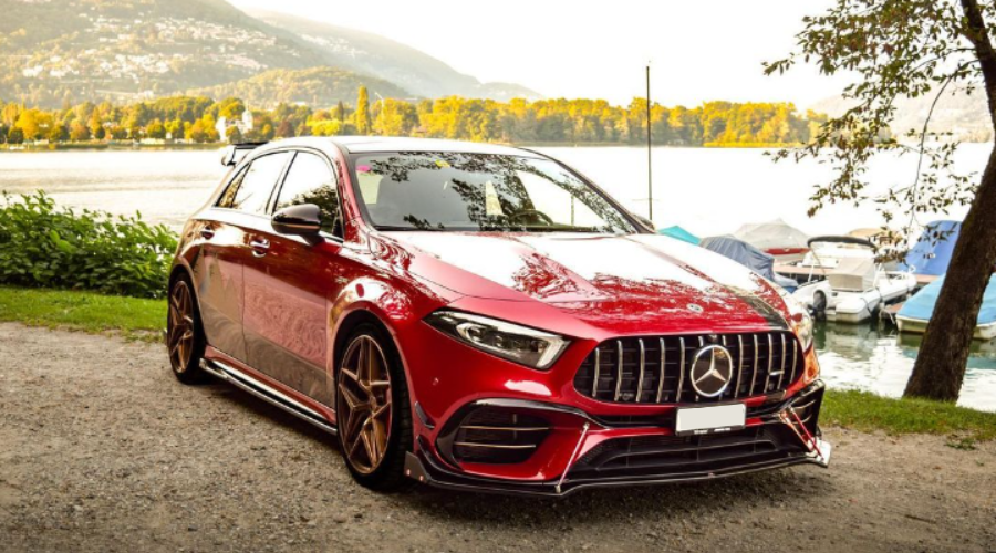 A red car with a prominent grille is parked on a gravel path by a lake. Boats and trees are in the background, with distant mountains under a clear sky.