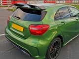 A green Mercedes-Benz hatchback is parked on a racetrack's asphalt surface. It's positioned near fencing and a grassy area, featuring a rear spoiler and tinted windows.