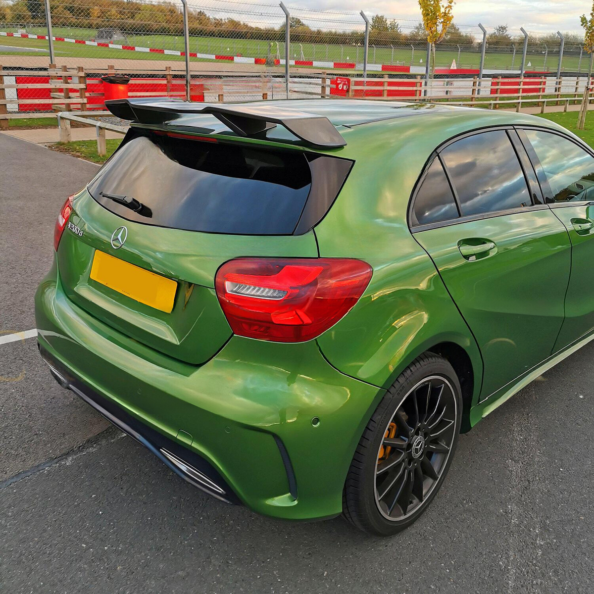 A green Mercedes-Benz hatchback is parked on a racetrack's asphalt surface. It's positioned near fencing and a grassy area, featuring a rear spoiler and tinted windows.