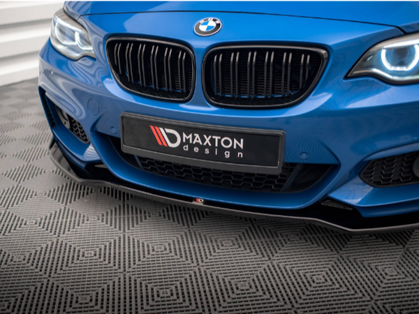 A blue BMW car features prominently with illuminated headlights on a patterned gray surface. The front license plate reads "MAXTON design." The setting is likely a showroom or garage.