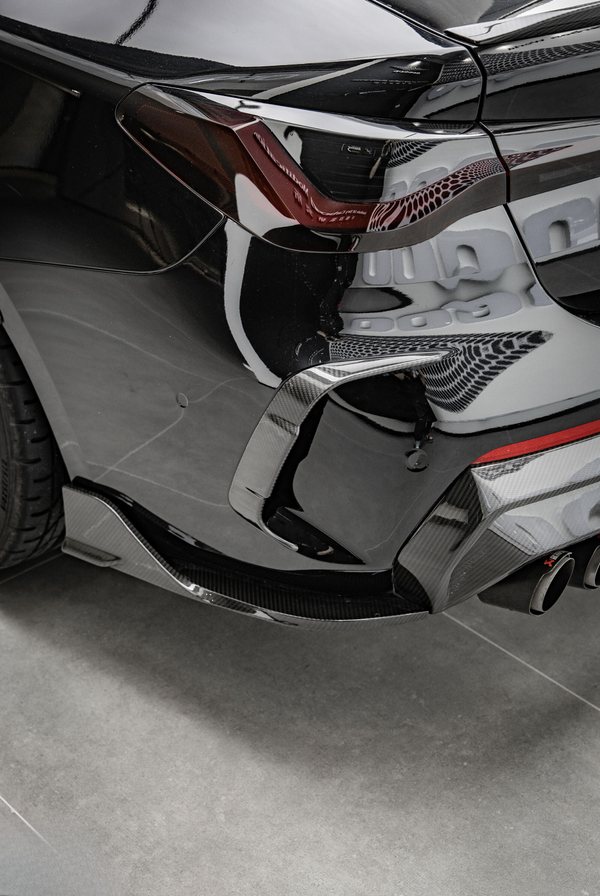 A sleek black car's rear corner, showcasing a tinted taillight and dual exhaust pipes, is parked indoors on a smooth gray floor.