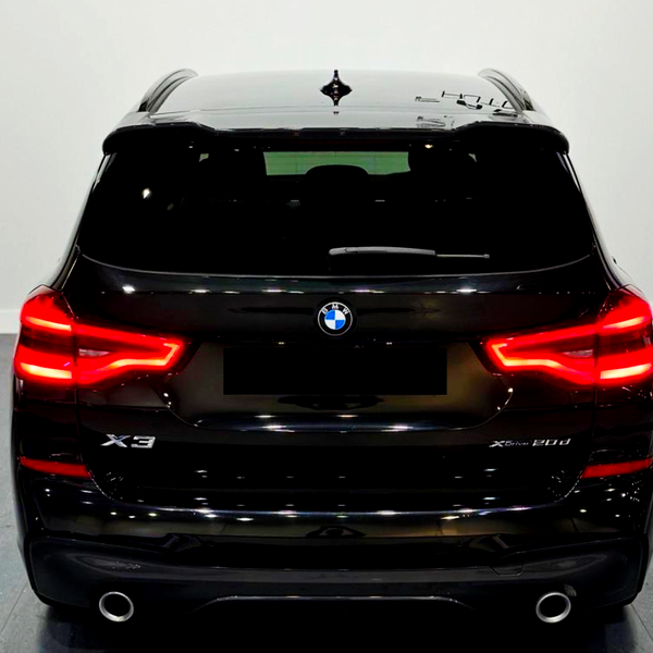 A black BMW X3 is parked indoors, with bright red taillights illuminated. The model designation "X3 xDrive 20d" appears on the back in a well-lit showroom.