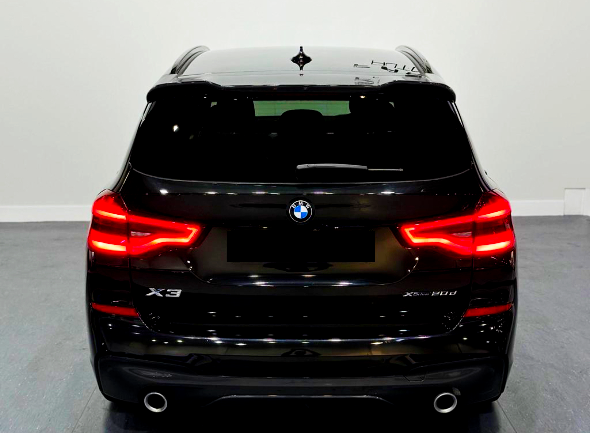 A black BMW X3 is parked indoors, with bright red taillights illuminated. The model designation "X3 xDrive 20d" appears on the back in a well-lit showroom.