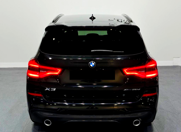 A black BMW X3 is parked indoors, with bright red taillights illuminated. The model designation "X3 xDrive 20d" appears on the back in a well-lit showroom.