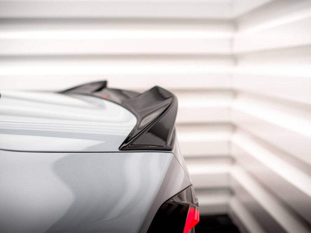 A car's rear spoiler is visible in close-up, featuring a glossy black finish. It is situated in a brightly lit, white-paneled room, enhancing its sleek design.
