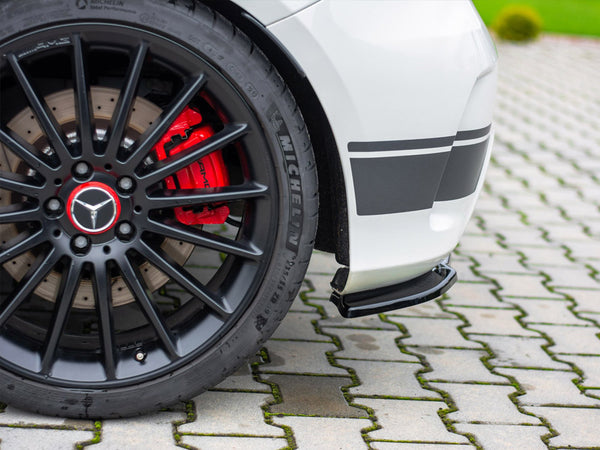 A black car wheel with a red brake caliper labeled "AMG" rests on a paved surface, adjacent to a white car body with black stripes. The tire reads "MICHELIN."