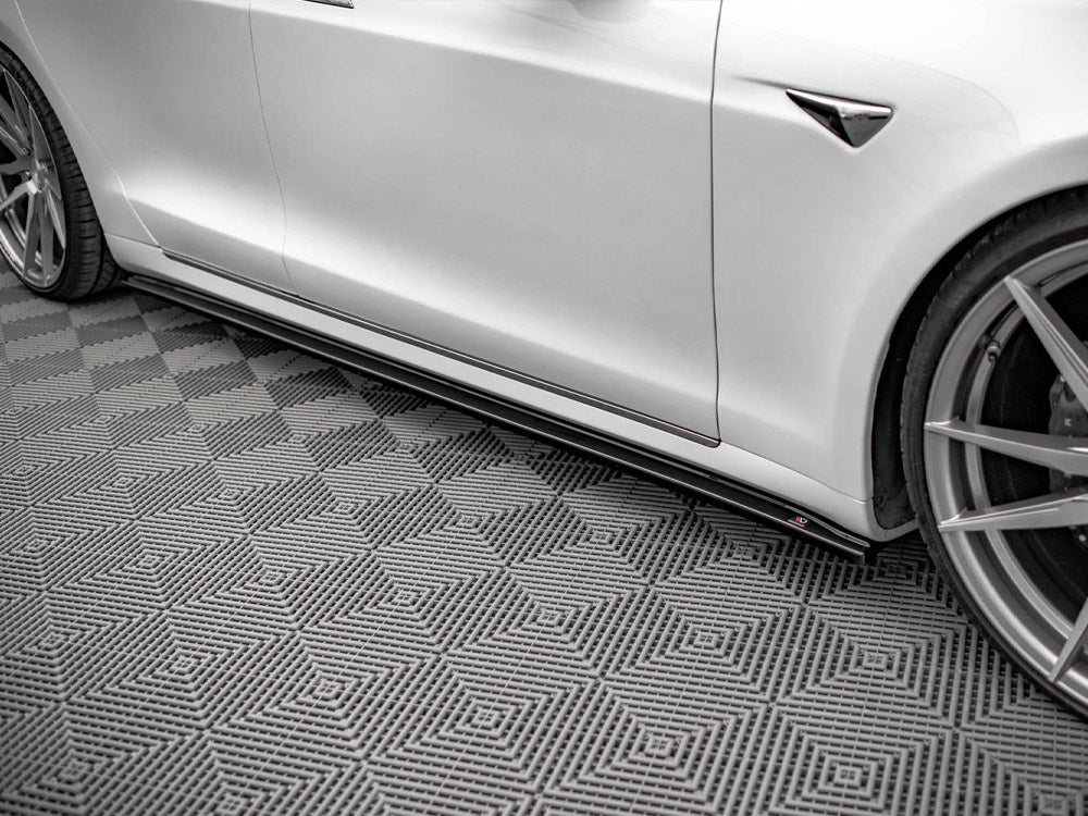 A white car with sleek, angular design rests stationary on a textured, diamond-patterned gray floor, showcasing its aerodynamic side skirts and alloy wheels in a well-lit indoor setting.
