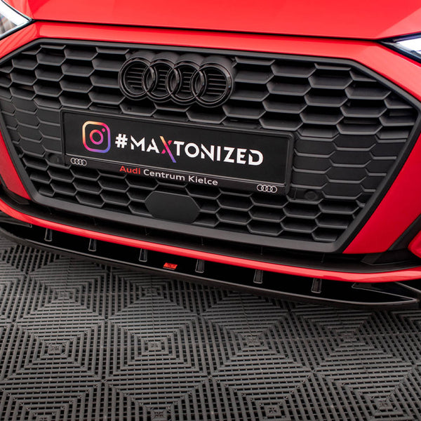 A red car's front grille dominates, with a black license plate reading "#MAXTONIZED Audi Centrum Kielce" against a textured black floor in a showroom setting.
