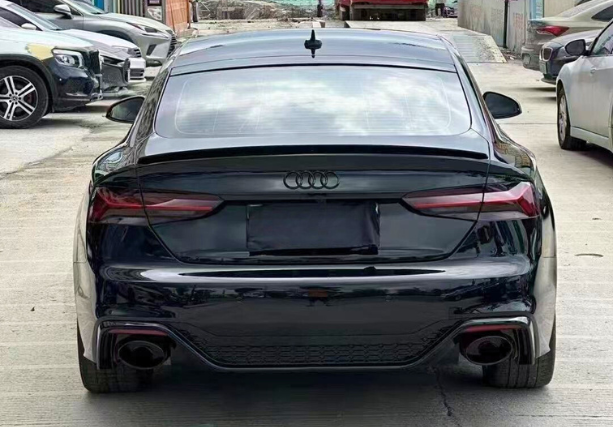 A black Audi car is parked on a concrete surface, surrounded by other vehicles in an industrial area.