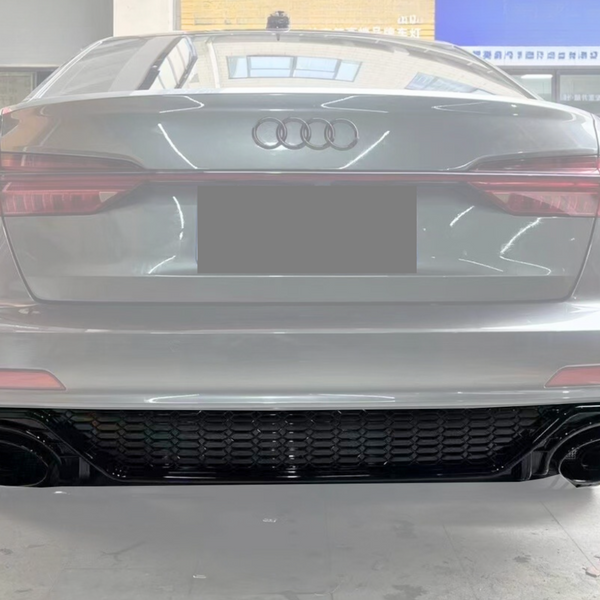 A silver Audi car is stationary inside a showroom, displaying its rear view with distinctive taillights and dual exhaust, parked among other cars under bright, reflective lighting.