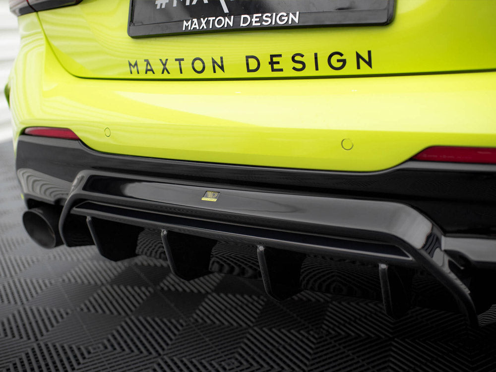 Bright yellow car features a black rear diffuser. "#MAXTON DESIGN" text is on the vehicle and license plate, parked on a patterned black floor.