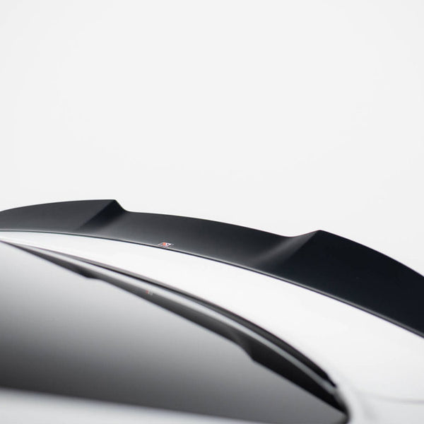 A black car spoiler is mounted on a sleek, white vehicle in a minimalistic setting. The background is a plain, light gray color, emphasizing the spoiler's contours.