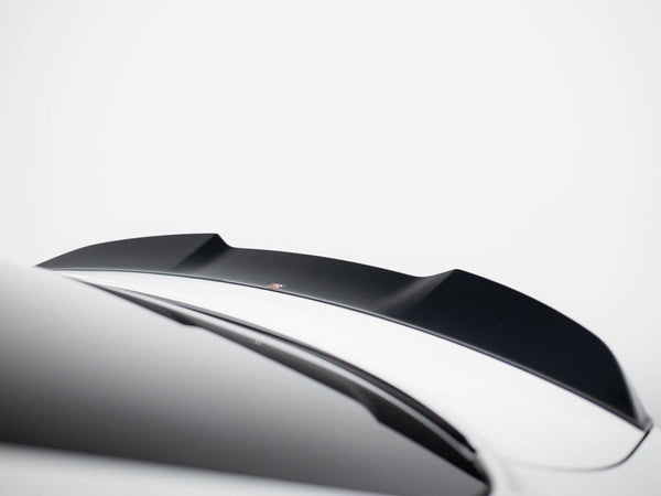 A black car spoiler is mounted on a sleek, white vehicle in a minimalistic setting. The background is a plain, light gray color, emphasizing the spoiler's contours.