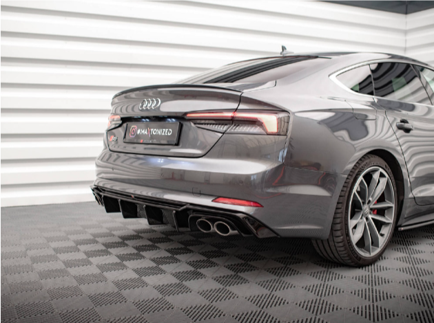 A sleek, gray Audi car stands stationary on a checkered floor. Its rear features dual exhausts and distinct LED taillights. The setting appears to be a well-lit indoor showroom. Text on the license plate reads "WRAPTONIZED."