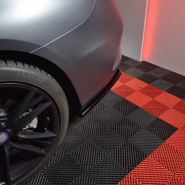 A car's rear wheel stands stationary on a checkered black and red floor in a well-lit room. The tire reads, "245/40 R 18 93 W M+S," and "OUTSIDE."