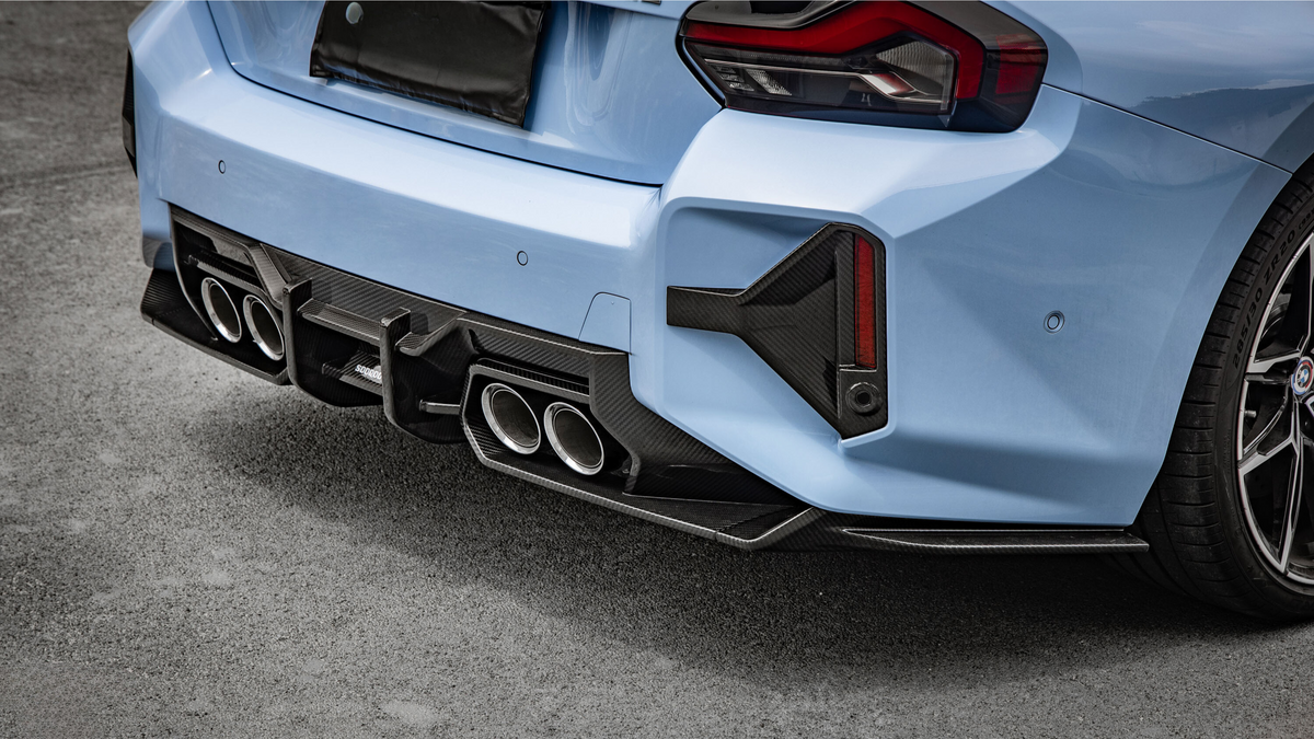 A light blue car with sporty dual exhaust pipes and a carbon fiber diffuser is parked on a paved surface. The rear taillights have an angular design.