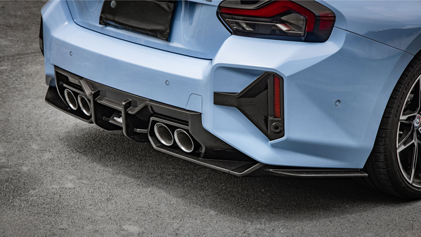 A light blue car with sporty dual exhaust pipes and a carbon fiber diffuser is parked on a paved surface. The rear taillights have an angular design.