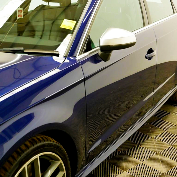 A shiny blue car is parked, showcasing a sleek design with a side mirror and a reflection on the side panel. It is indoors on a patterned floor. There is a yellow sticker with "PASSED" on the windshield.