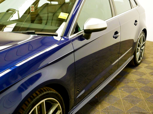 A shiny blue car is parked, showcasing a sleek design with a side mirror and a reflection on the side panel. It is indoors on a patterned floor. There is a yellow sticker with "PASSED" on the windshield.