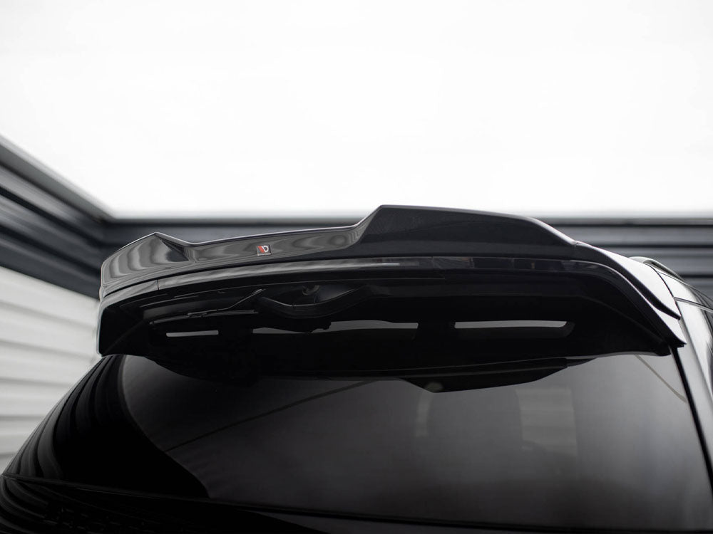 A shiny black car's rear spoiler is raised, highlighting its glossy finish. In a dimly lit garage setting, a small red and black "V" logo is visible on the spoiler.