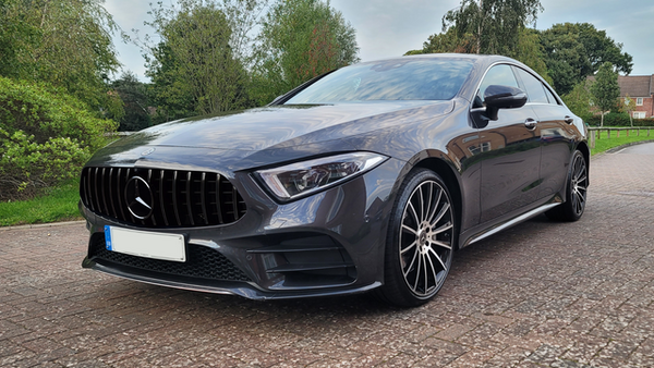 A gray Mercedes-Benz sedan is parked on a brick driveway, surrounded by green trees and a residential area. The car has sleek lines and prominent black wheels.