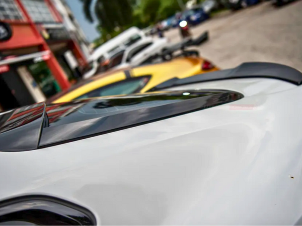 A white car's rear end with a spoiler is parked on a street, surrounded by other vehicles and buildings. The street has a relaxed urban environment.