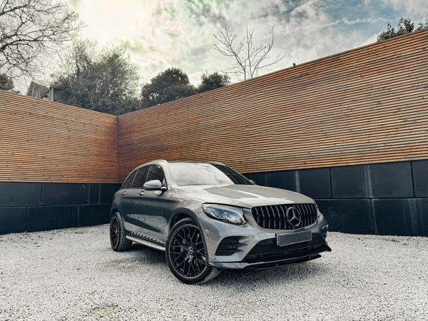 A silver SUV is parked on a gravel surface. The vehicle is positioned in front of a wooden fence, with trees visible in the background under a cloudy sky.