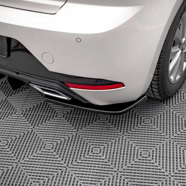 The rear of a silver car with a visible tire, exhaust pipe, and red reflector rests on a textured gray, grid-patterned surface. Partial signage reads "XTON Design."