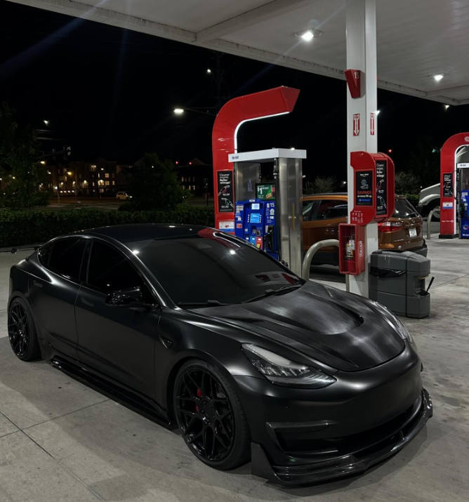 A sleek black car is parked under bright lights at a gas station, near fuel pumps and trash bins, with a nighttime backdrop of trees and distant buildings.