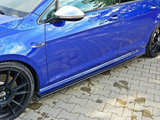 A blue car is parked on a wet, paved surface. The reflection of trees can be seen on the car's shiny surface, and black rims are visible on the wheels.