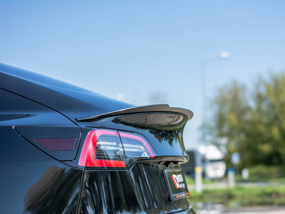 A sleek black car with a prominent rear spoiler is parked outdoors, with clear taillights visible. Trees and sky form the background, providing a serene setting.