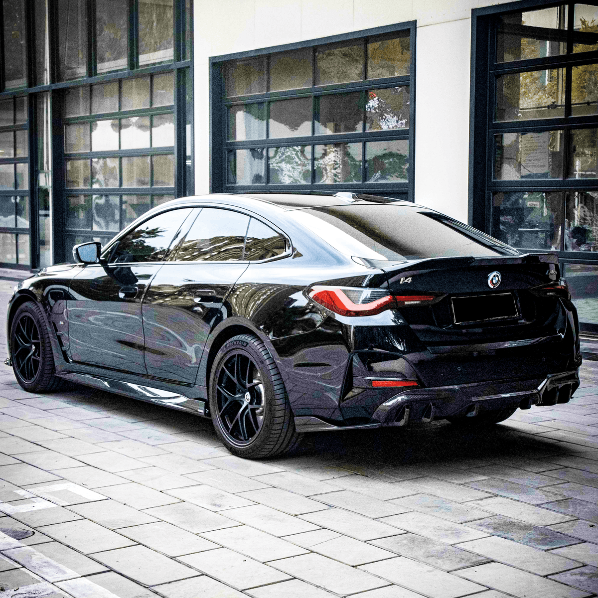 A sleek black BMW i4 is parked on a tiled urban street, reflecting nearby buildings in its shiny surface, against a backdrop of large windows.