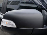 A black car side mirror with an integrated turn signal is attached to a vehicle's door, reflecting an urban setting.