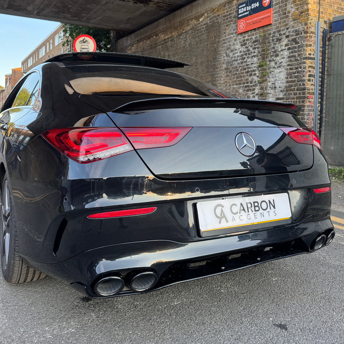 Car with a sleek black exterior parked under a bridge, displaying a "Carbon Accents" license plate. Red taillights glow, while a sign above warns of restricted vehicle height.