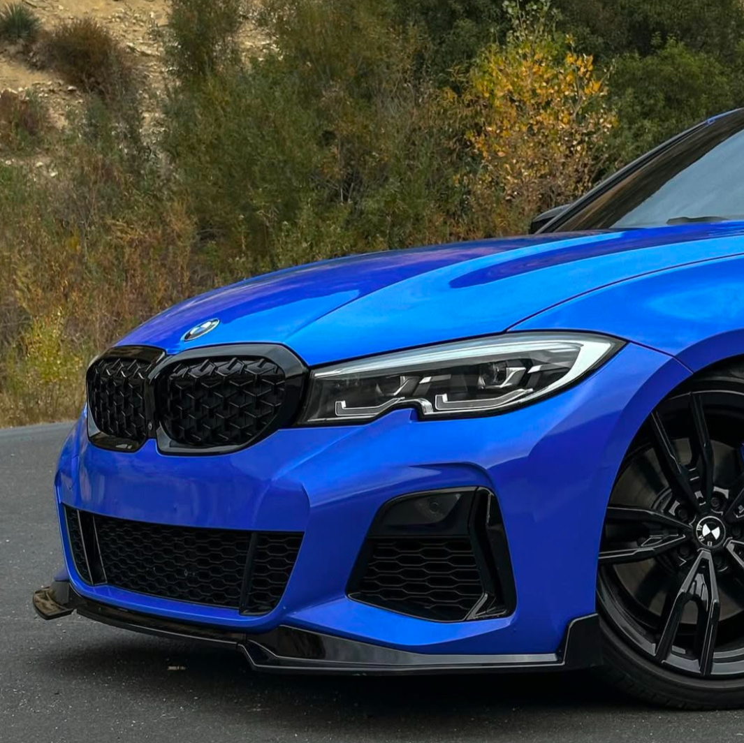 A blue car with a prominent grille and sleek headlights is parked on a road, surrounded by trees and shrubs. The shiny black wheel is partially visible.