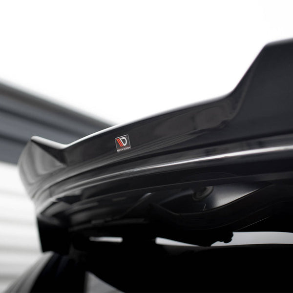 A black car spoiler features a "Vortex Generator" sticker, mounted on the vehicle's trunk. The background includes blurred, horizontal lines of a light-colored structure, suggesting an industrial environment.
