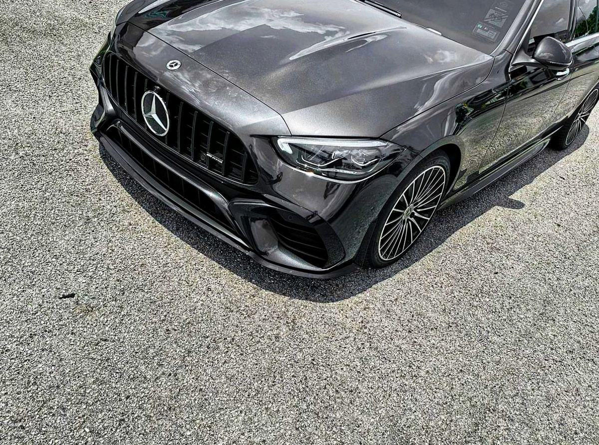 A sleek, dark-colored Mercedes car is parked on a gravel surface, highlighting its prominent grille and intricate wheel design under a partly cloudy sky.