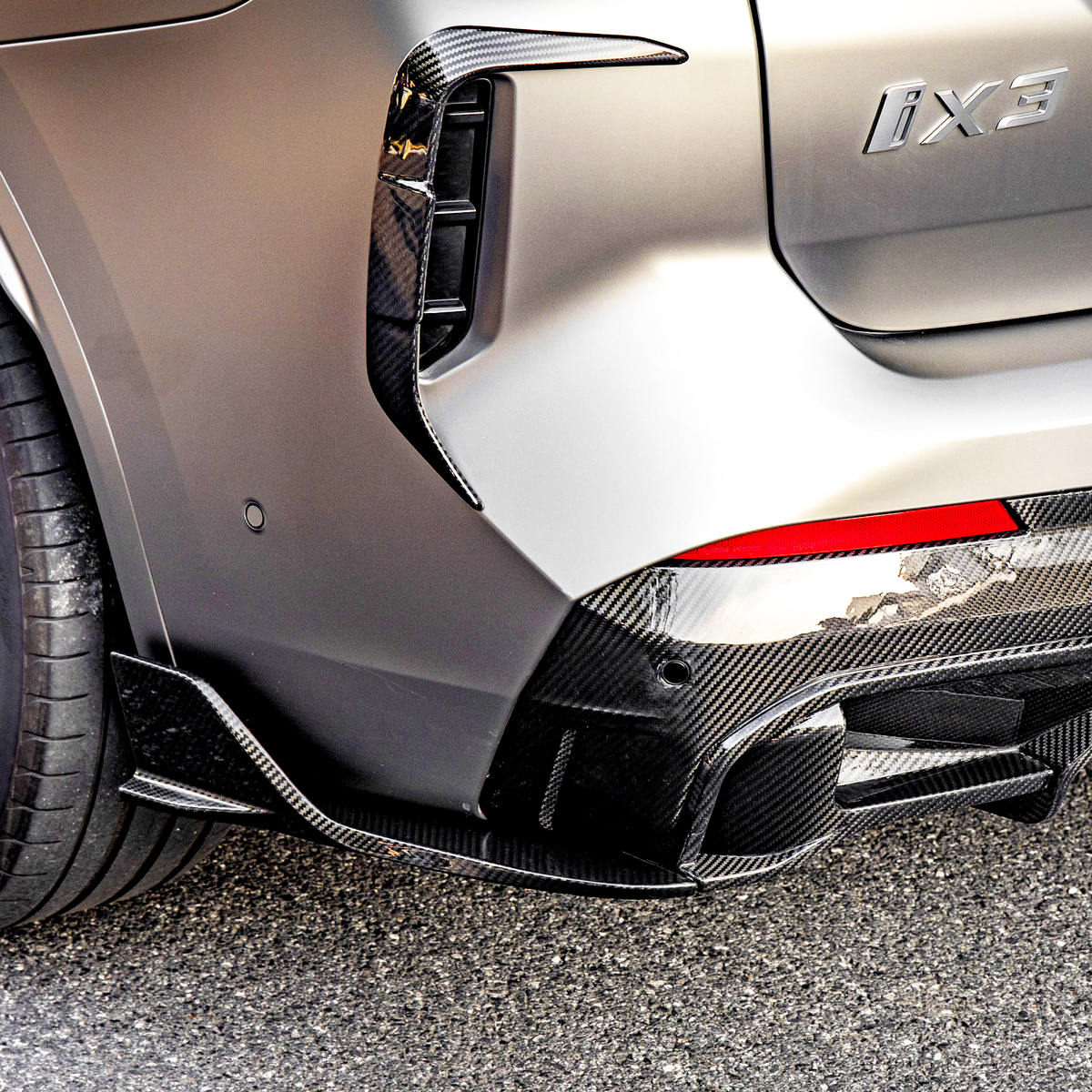A car's rear section with a silver body and carbon fiber trim showcases a sleek tire design and red taillight. The badge reads "iX3." It's parked on asphalt.
