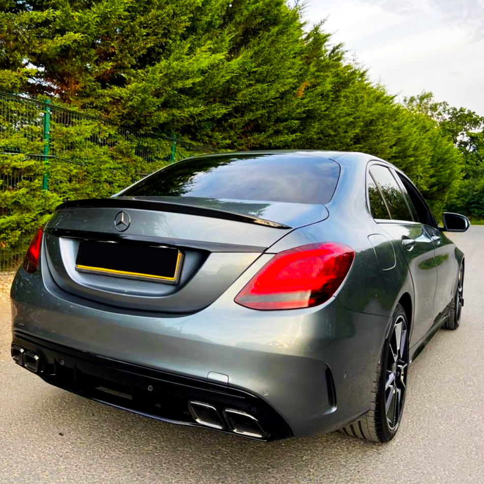 A silver Mercedes-Benz sedan is parked on the street, facing away. It's alongside a tall, green hedge under a cloudy sky, with no visible text.