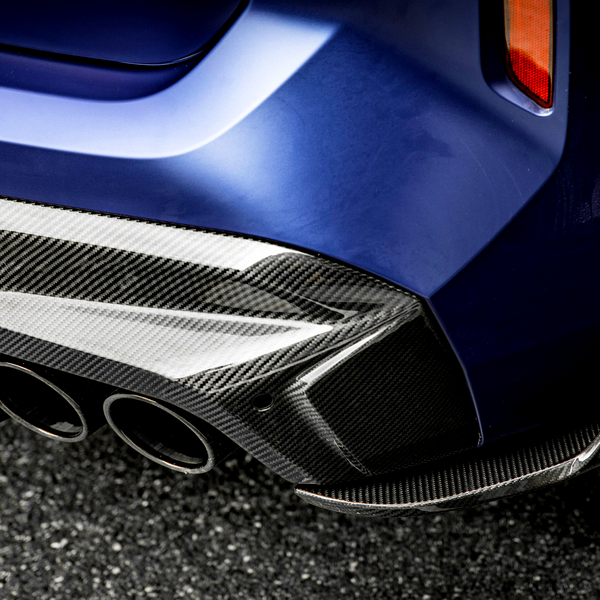 A car's blue rear bumper features a glossy carbon fiber diffuser with three exhaust pipes, parked on an asphalt surface. The setting suggests a modern automotive design.