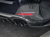 A car's rear bumper features dual exhaust pipes and a carbon fiber diffuser. It's parked on asphalt with a visible red reflector strip.